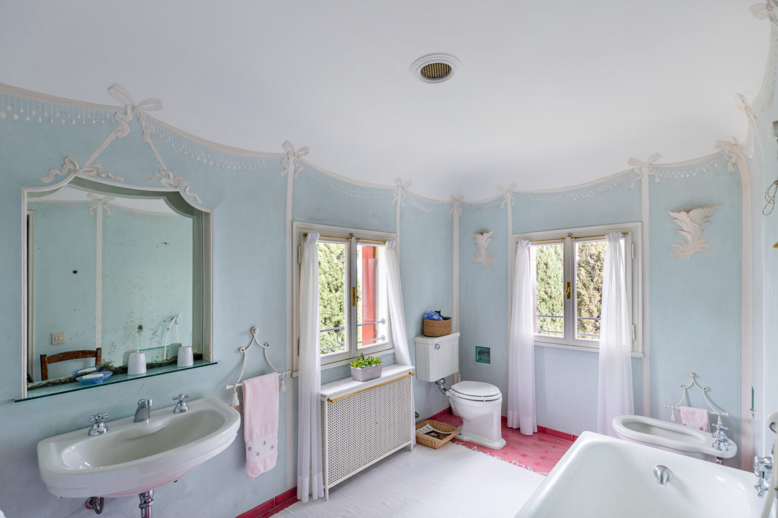 Bright and airy bathroom featuring a light blue color scheme, decorative molding, and elegant fixtures. The space includes a vintage bathtub, a pedestal sink, and a white toilet, complemented by natural light from windows adorned with sheer curtains. A small potted plant and decorative elements enhance the charming atmosphere. | Only on SopranoVillas