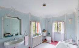 Bright and airy bathroom featuring a light blue color scheme, decorative molding, and elegant fixtures. The space includes a vintage bathtub, a pedestal sink, and a white toilet, complemented by natural light from windows adorned with sheer curtains. A small potted plant and decorative elements enhance the charming atmosphere.