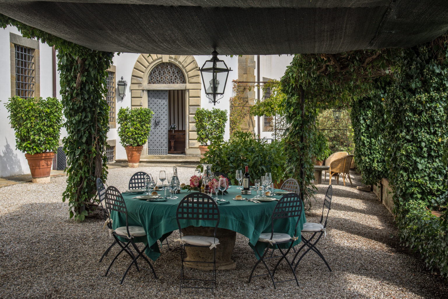 Outdoor dining setup in a lush garden with a green tablecloth, surrounded by potted plants and ivy-covered walls, creating an inviting atmosphere for gatherings. | Only on SopranoVillas