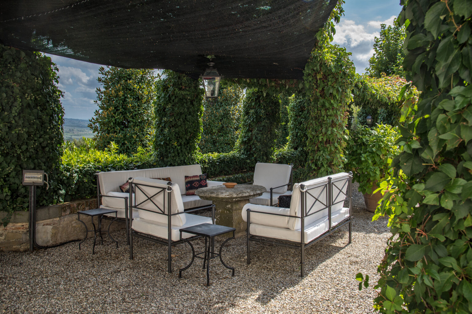 Cozy outdoor seating area with white upholstered chairs and a stone table, surrounded by lush greenery and a shaded canopy, perfect for relaxation and social gatherings. | Only on SopranoVillas