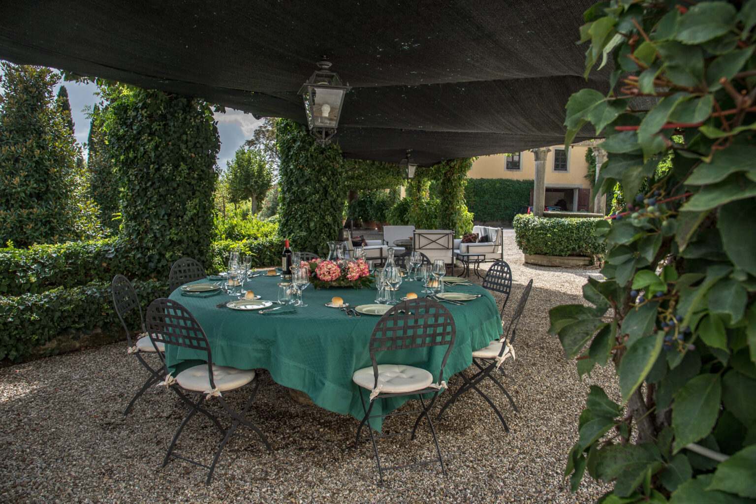 Outdoor dining setup under a shaded pergola with a green tablecloth, surrounded by lush greenery and decorative lanterns, creating an inviting atmosphere for gatherings or events. | Only on SopranoVillas