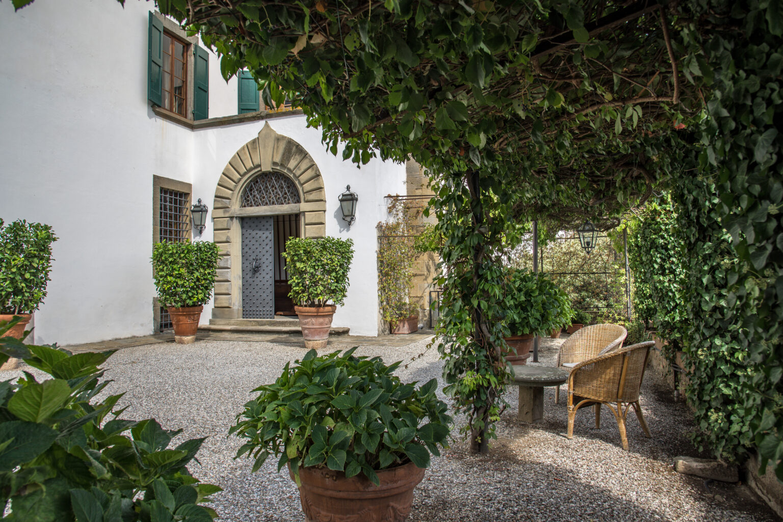 Charming entrance of a historic villa surrounded by lush greenery and potted plants, featuring a gravel pathway and inviting outdoor seating area. | Only on SopranoVillas