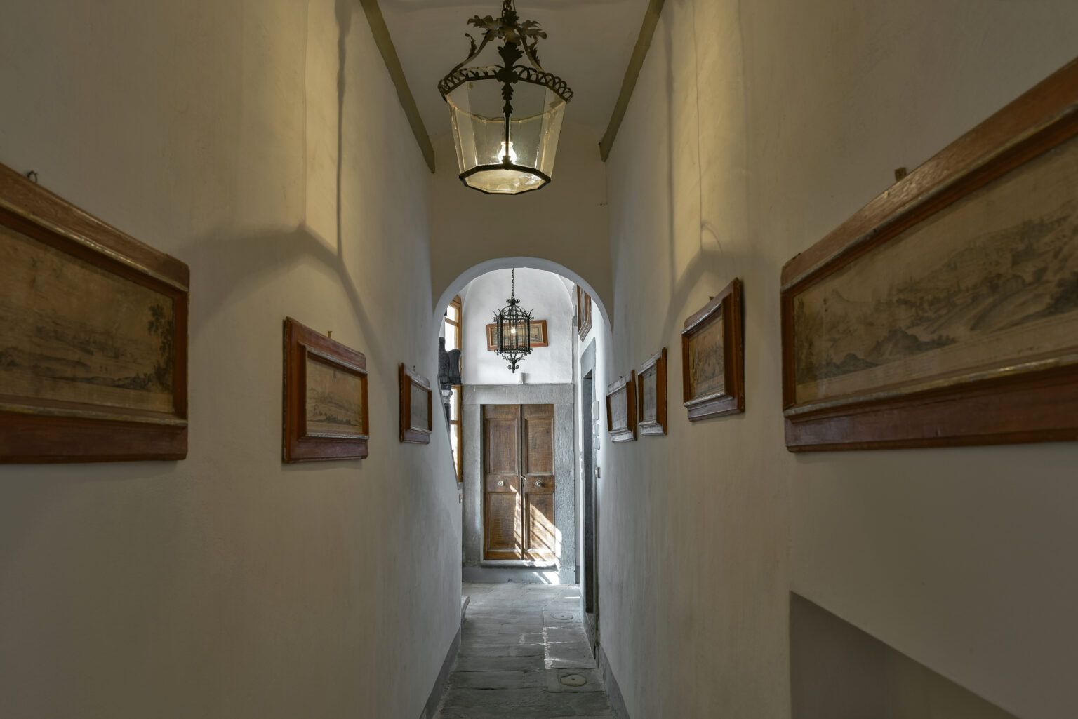 Narrow hallway featuring framed artwork on white walls, illuminated by a decorative lantern, leading to a wooden door and natural light from the outside. | Only on SopranoVillas