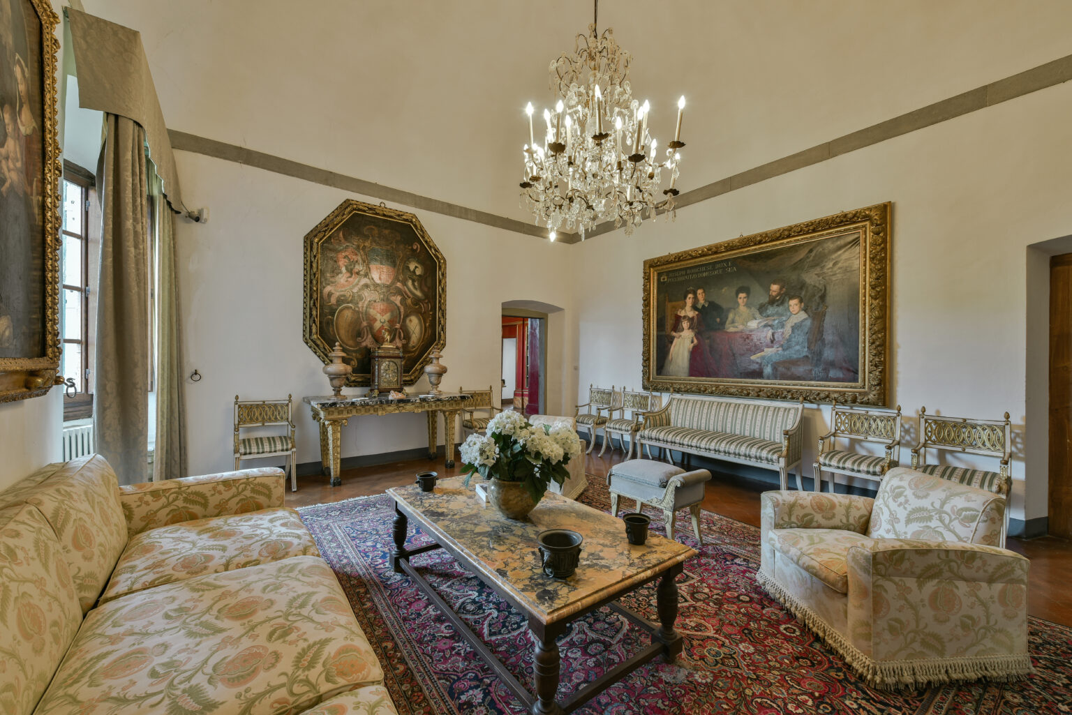 Elegant living room featuring a chandelier, ornate paintings, and vintage furniture, including a floral sofa and a decorative coffee table with a vase of flowers. The room is adorned with rich textures and colors, creating a luxurious atmosphere. | Only on SopranoVillas