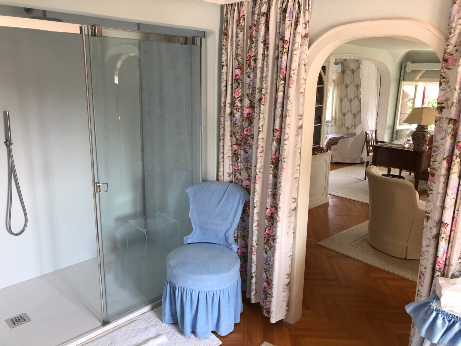 Bathroom interior featuring a glass shower enclosure, a blue upholstered chair, and floral curtains, with a view into a cozy living area that includes a desk and decorative elements. | Only on SopranoVillas