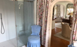 Bathroom interior featuring a glass shower enclosure, a blue upholstered chair, and floral curtains, with a view into a cozy living area that includes a desk and decorative elements.