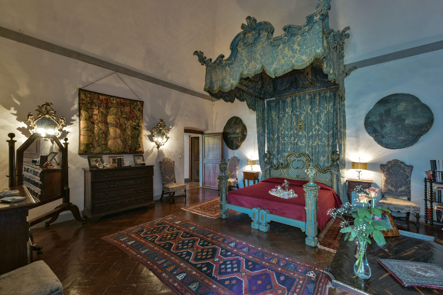 Luxurious vintage bedroom featuring an ornate canopy bed with intricate carvings, rich red bedding, antique furniture, and decorative wall art. The room is illuminated by soft lighting, creating an elegant and inviting atmosphere. | Only on SopranoVillas