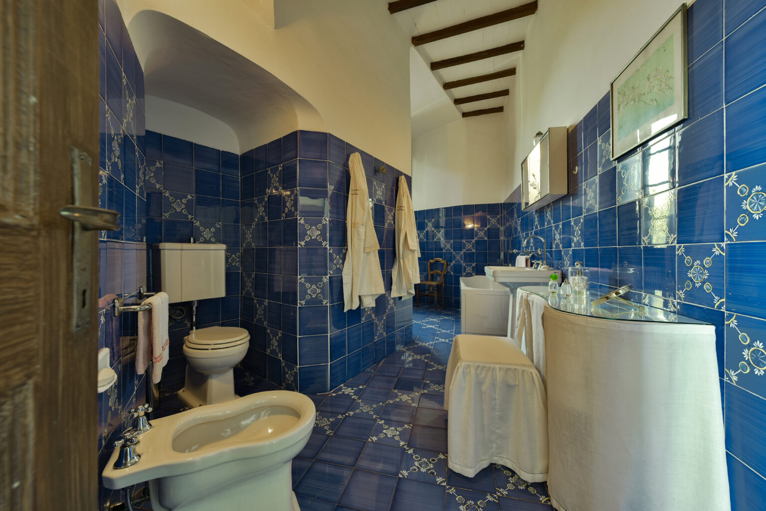 Brightly colored blue tiled bathroom featuring a toilet, sink, and elegant decor. The space is complemented by hanging towels and a stylish design, showcasing a blend of functionality and aesthetic appeal. Ideal for home or hospitality settings. | Only on SopranoVillas