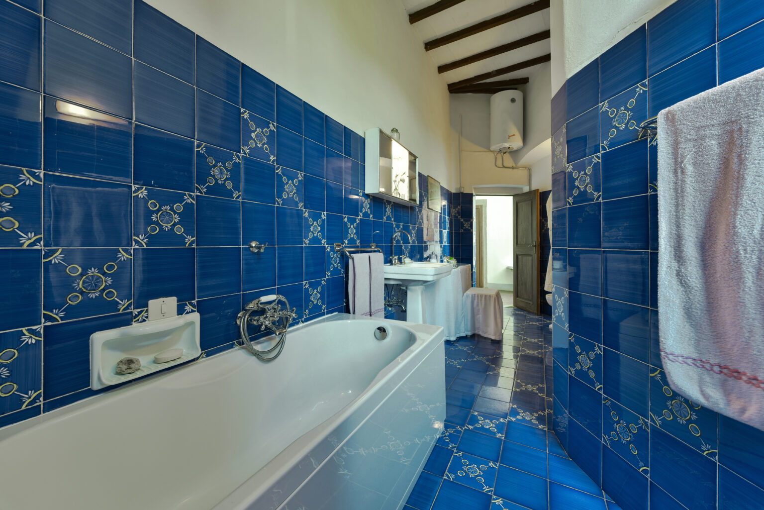 Modern bathroom featuring vibrant blue tiled walls, a bathtub, and a stylish sink area, accented by wooden beams and decorative patterns. | Only on SopranoVillas