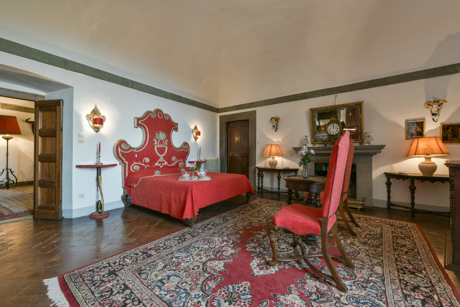 Elegant bedroom featuring a red-themed decor with a vintage bed, ornate headboard, and classic furnishings. The room is accented by a large area rug, decorative lamps, and antique-style furniture, creating a warm and inviting atmosphere. | Only on SopranoVillas