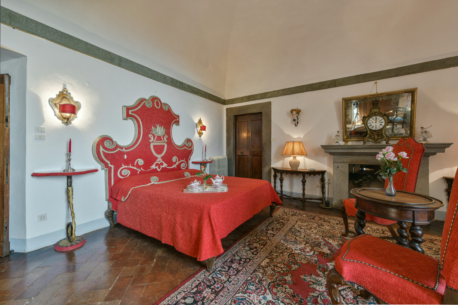 Elegant bedroom featuring a red upholstered bed with intricate detailing, matching decor, hardwood flooring, and a decorative fireplace. The room is adorned with stylish lighting and classic furnishings, creating a cozy and luxurious atmosphere. | Only on SopranoVillas