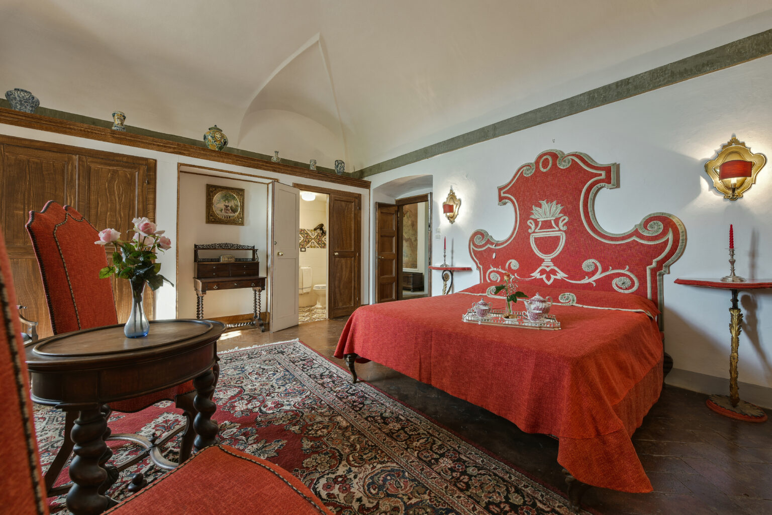Luxurious bedroom featuring a red upholstered bed with intricate design, vintage wooden furniture, and elegant decor, set in a spacious, well-lit room with arched ceilings and a rich area rug. | Only on SopranoVillas