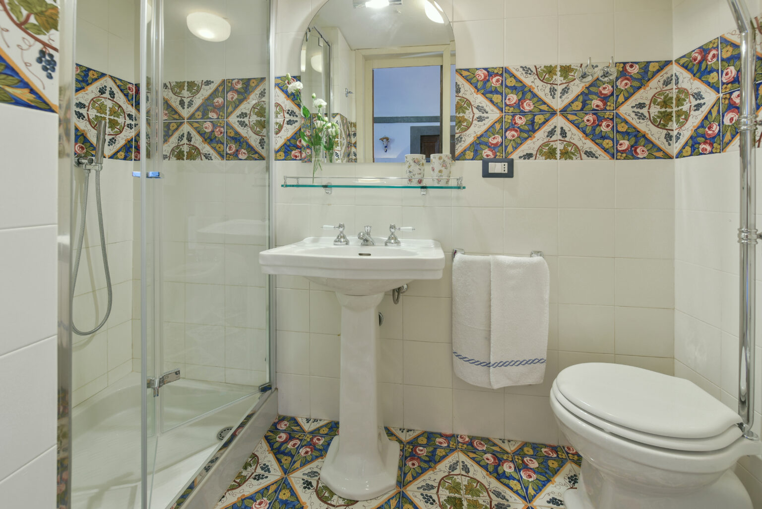 Modern bathroom featuring a glass shower, elegant sink with two faucets, and decorative tiled walls. The space includes a white toilet and a neatly arranged towel, showcasing a stylish and functional design. | Only on SopranoVillas