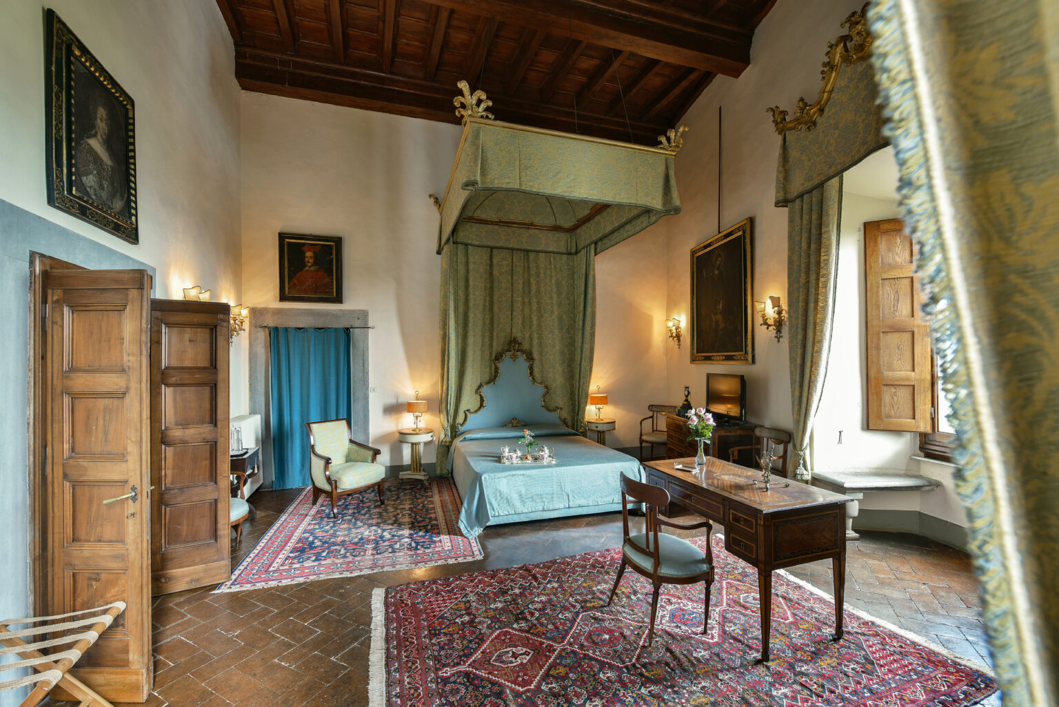 Luxurious bedroom interior featuring a four-poster canopy bed, elegant furnishings, and rich wooden beams. The room is adorned with classic artwork, decorative curtains, and a vintage writing desk, creating a serene and opulent atmosphere. | Only on SopranoVillas