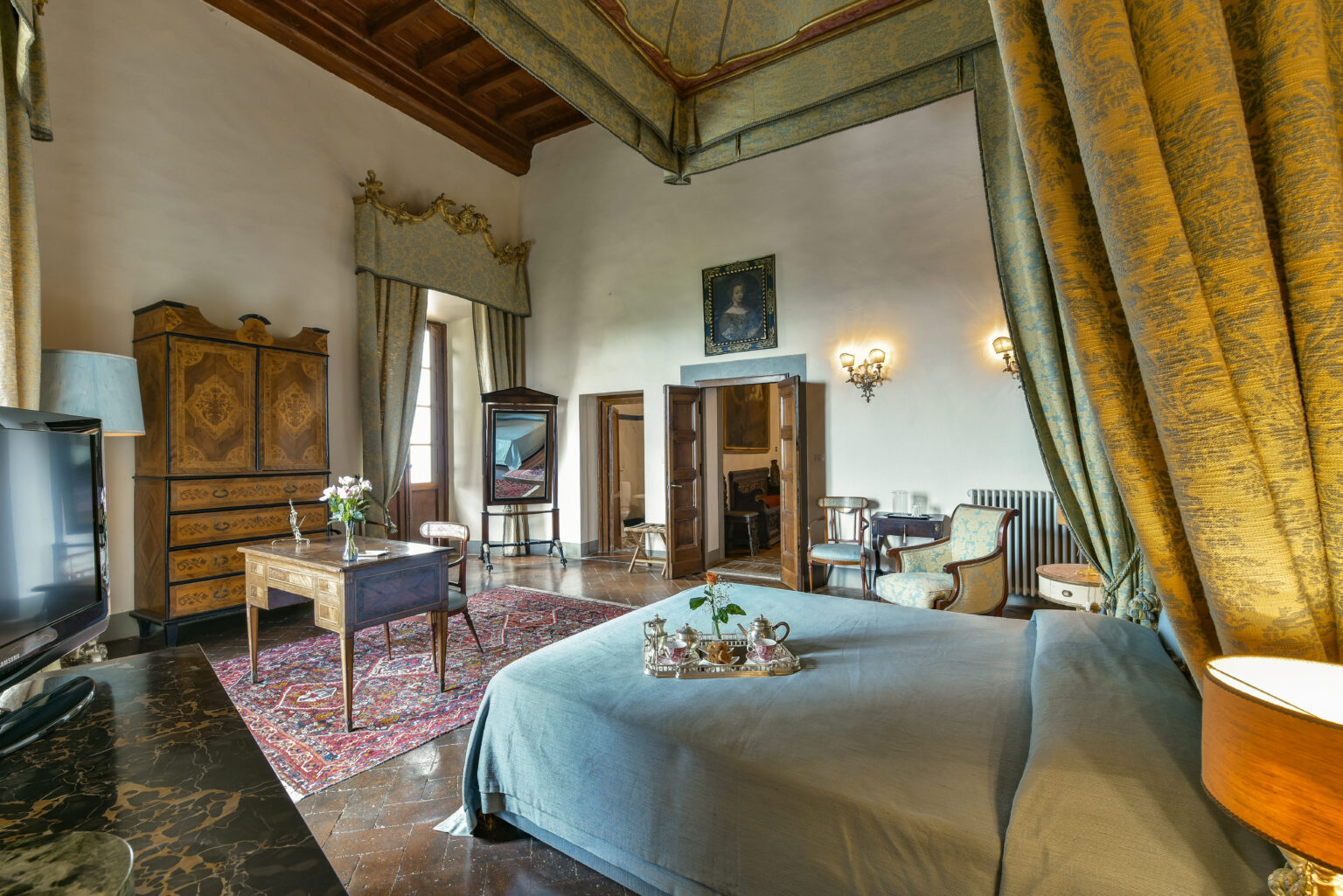 Luxurious hotel room featuring a spacious layout with a comfortable bed, stylish furnishings, elegant drapes, and a vintage wardrobe. The room is adorned with decorative elements, including a rug and artwork, creating a warm and inviting atmosphere. | Only on SopranoVillas