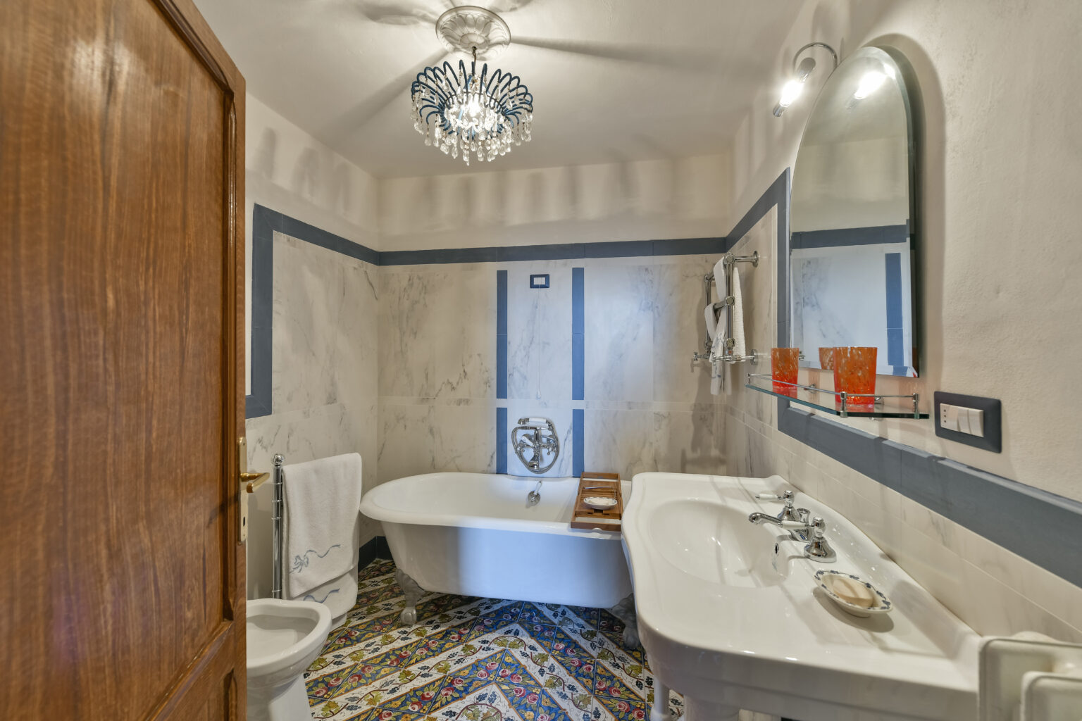 Elegant bathroom featuring a freestanding bathtub, modern sink, and decorative tile floor. The space is illuminated by a stylish chandelier and includes a large mirror, creating a bright and inviting atmosphere. | Only on SopranoVillas