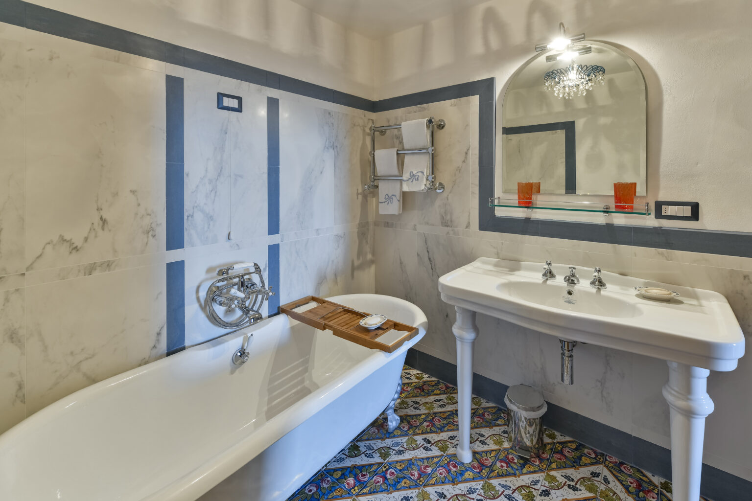 Elegant bathroom featuring a vintage freestanding bathtub, a stylish sink with ornate fixtures, and a decorative mirror. The walls are adorned with a subtle marble finish, complemented by colorful patterned tiles on the floor. Ideal for relaxation and luxury. | Only on SopranoVillas