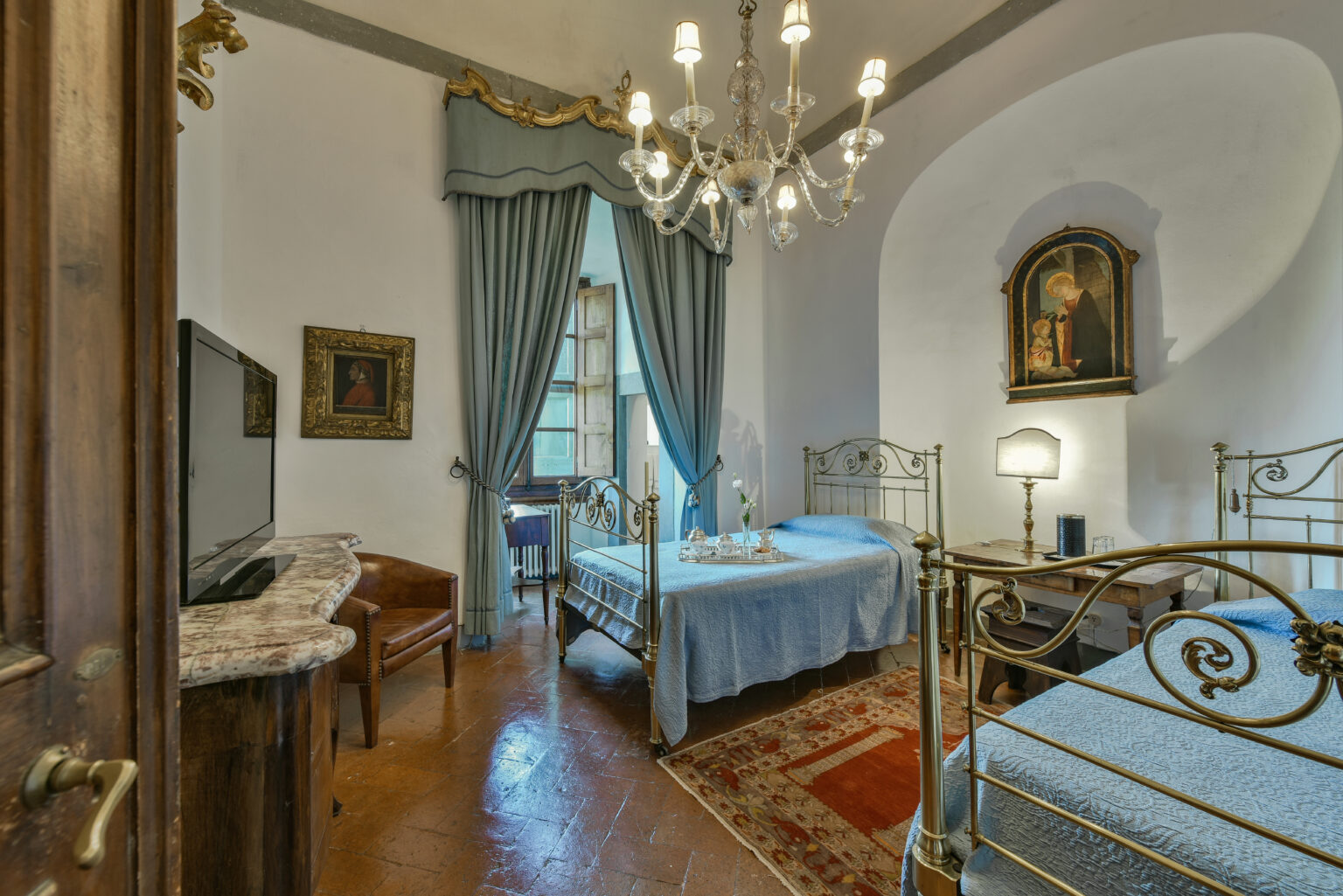 Elegant bedroom interior featuring two twin beds with blue bedding, a vintage chandelier, and classic decor. Natural light streams in through large windows adorned with blue curtains, complemented by a wooden furniture piece and an artwork on the wall. Ideal for a cozy and stylish stay. | Only on SopranoVillas
