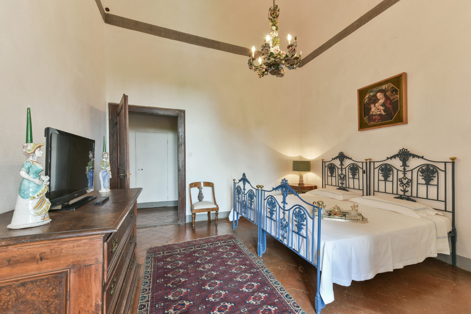 Cozy bedroom featuring a vintage wrought iron bed, wooden dresser, and a chandelier. The room is adorned with a decorative painting and a traditional area rug, creating an inviting atmosphere. | Only on SopranoVillas