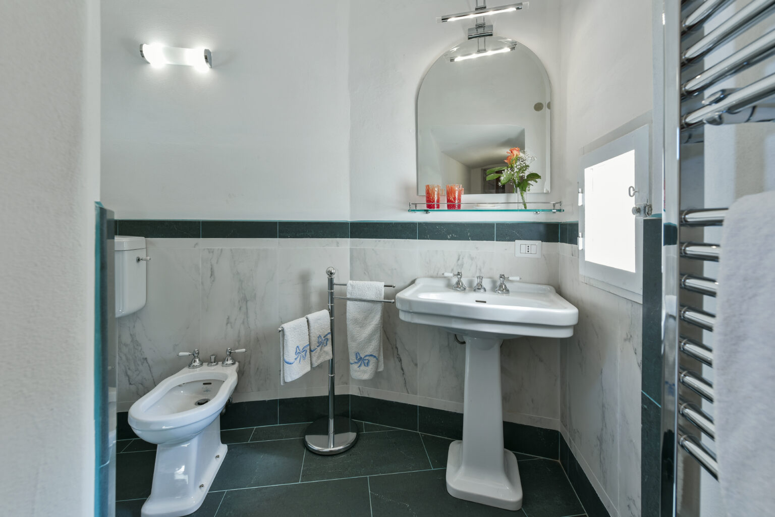 Modern bathroom interior featuring a white sink, toilet, and stylish mirror. The space is well-lit with natural light, accented by green tiles and a towel rack. Decorative plants add a touch of freshness. Ideal for showcasing contemporary bathroom design. | Only on SopranoVillas