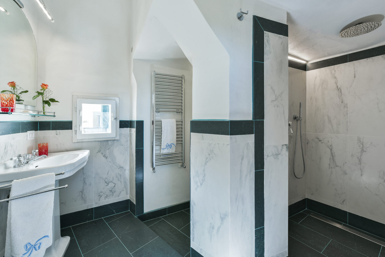 Modern bathroom featuring a sleek design with marble walls, a stylish sink, and a spacious shower area. The room is well-lit with natural light coming through a window, and decorative elements include flowers and a neatly arranged towel. | Only on SopranoVillas