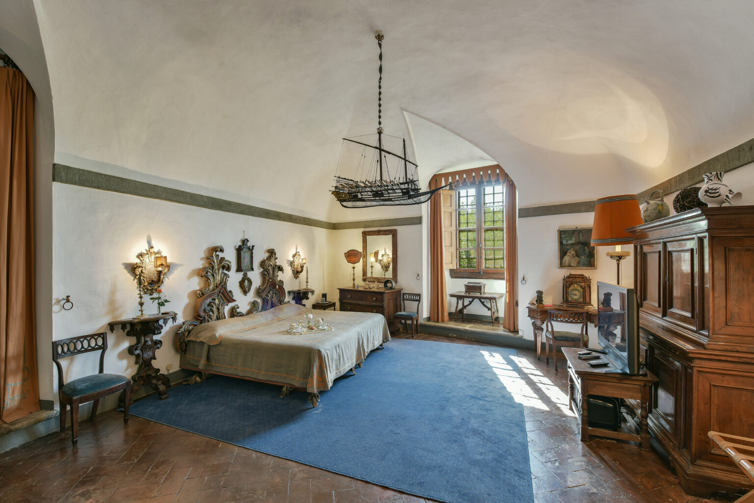 Elegant bedroom featuring a large bed with ornate headboard, vintage furniture, and a unique ship model hanging from the ceiling. The room has natural light streaming in from a large window, complemented by decorative wall sconces and rich textures throughout. | Only on SopranoVillas