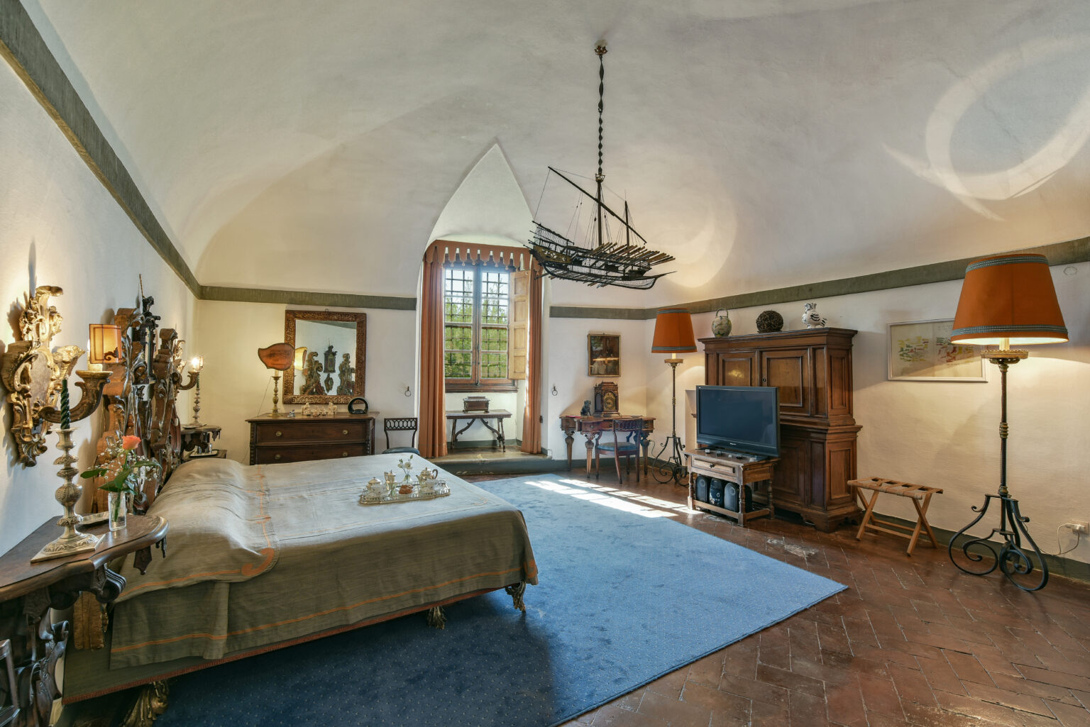 Spacious and elegantly designed bedroom featuring a vintage wooden bed, decorative wall sconces, and a unique ship-shaped chandelier. The room includes a cozy seating area, a wooden dresser, and a flat-screen TV, with natural light streaming through large windows adorned with orange curtains. The warm color palette and antique furnishings create a charming atmosphere. | Only on SopranoVillas