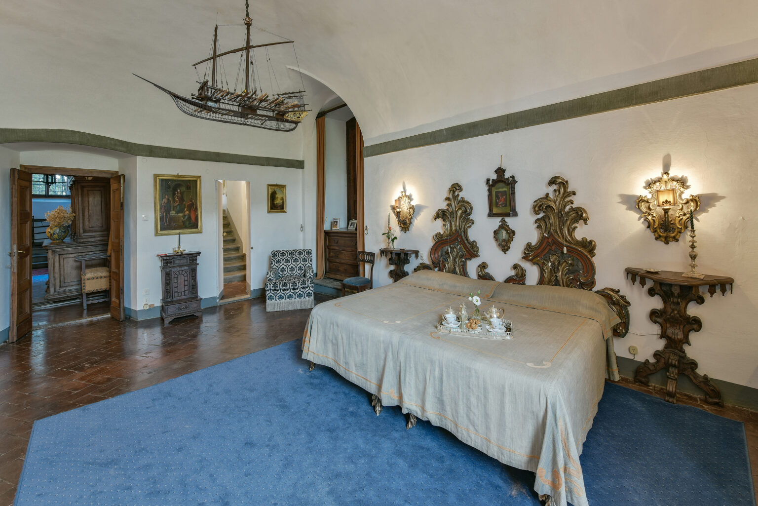 Spacious and elegantly decorated bedroom featuring a large bed with ornate headboards, a blue carpet, and vintage furnishings. The room includes a decorative ship model hanging from the ceiling, antique lighting fixtures, and artwork on the walls, creating a charming and inviting atmosphere. | Only on SopranoVillas