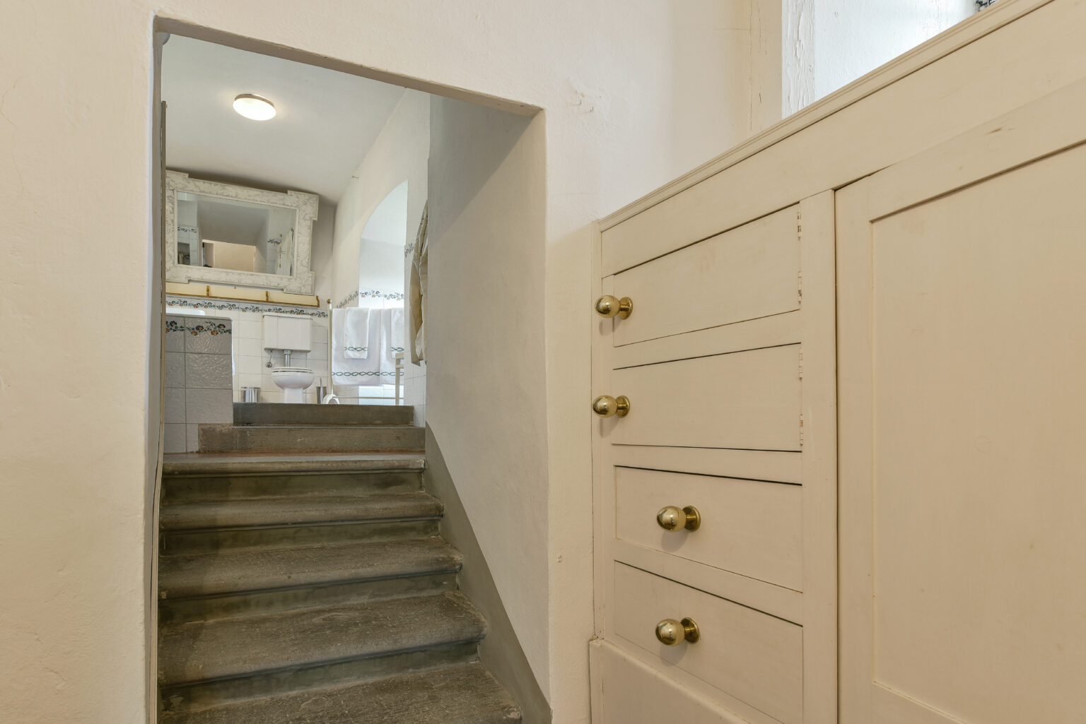 Interior view of a staircase leading to a bathroom, featuring white walls, a wooden cabinet with brass knobs, and a glimpse of tiled bathroom decor in the background. | Only on SopranoVillas