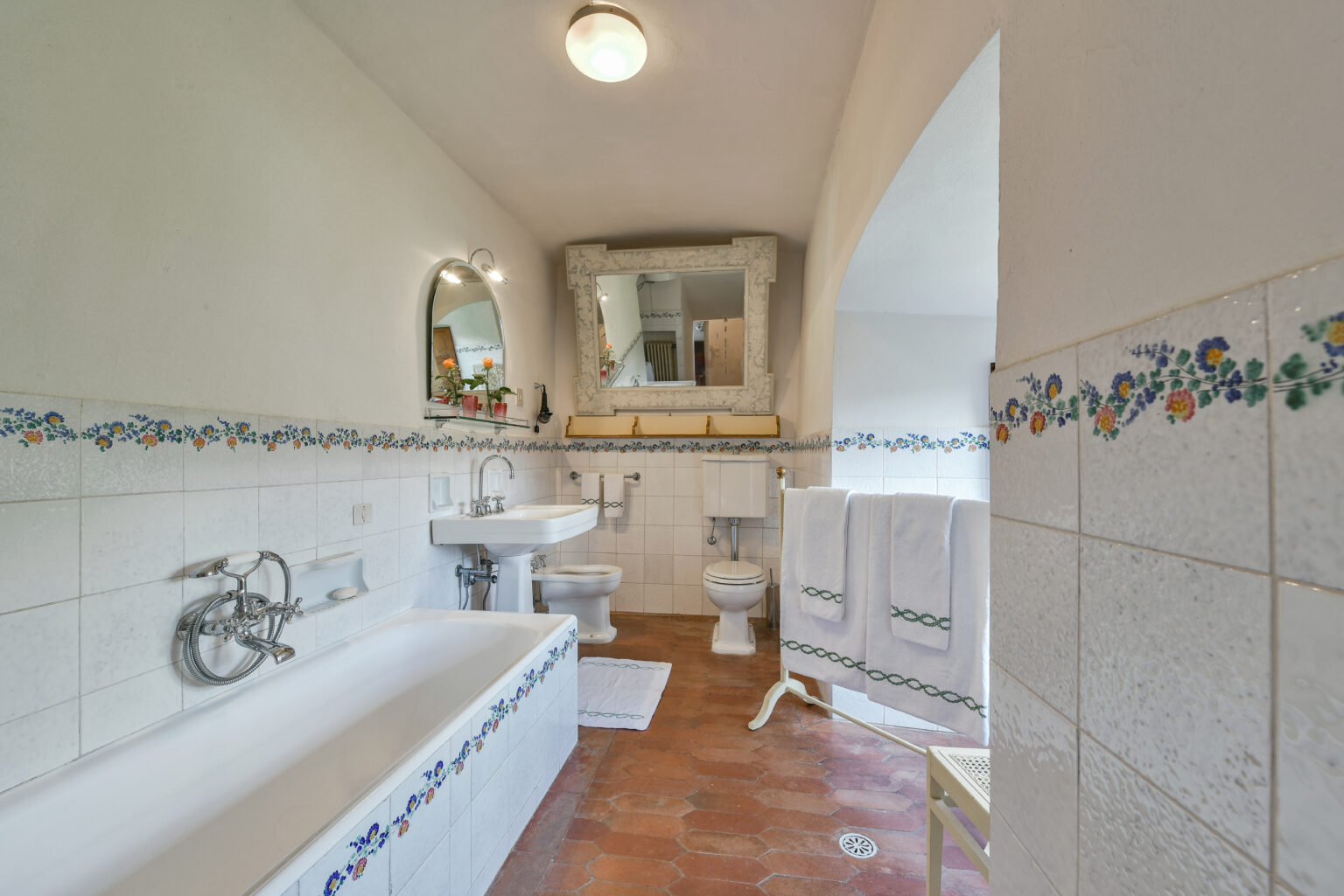 Bright and spacious bathroom featuring a white bathtub, modern sink, and toilet. The room is adorned with floral-patterned tiles and a large mirror, complemented by wooden accents and neatly arranged towels. Natural light enhances the inviting atmosphere. | Only on SopranoVillas