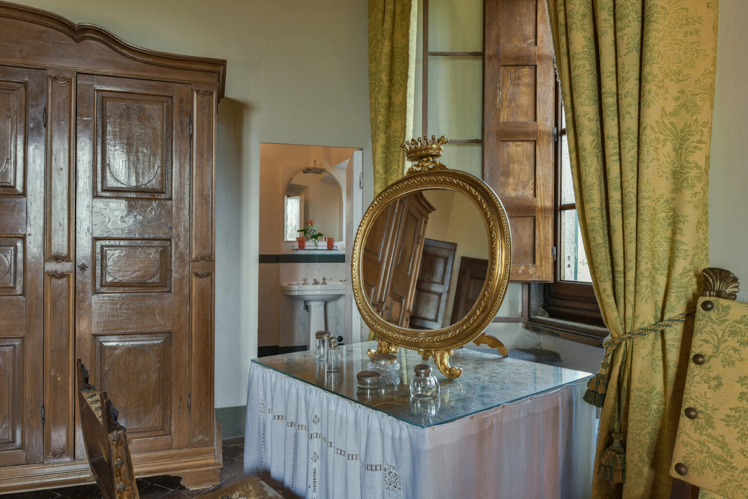 Elegant bedroom interior featuring a vintage wooden wardrobe, a round mirror with an ornate frame on a glass table, and lush green curtains. The setting exudes classic charm and sophistication. | Only on SopranoVillas
