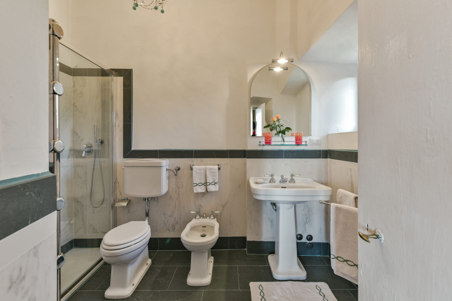 Modern bathroom featuring a glass shower, white sink, bidet, and toilet, complemented by dark tile flooring and decorative plants on the countertop. | Only on SopranoVillas