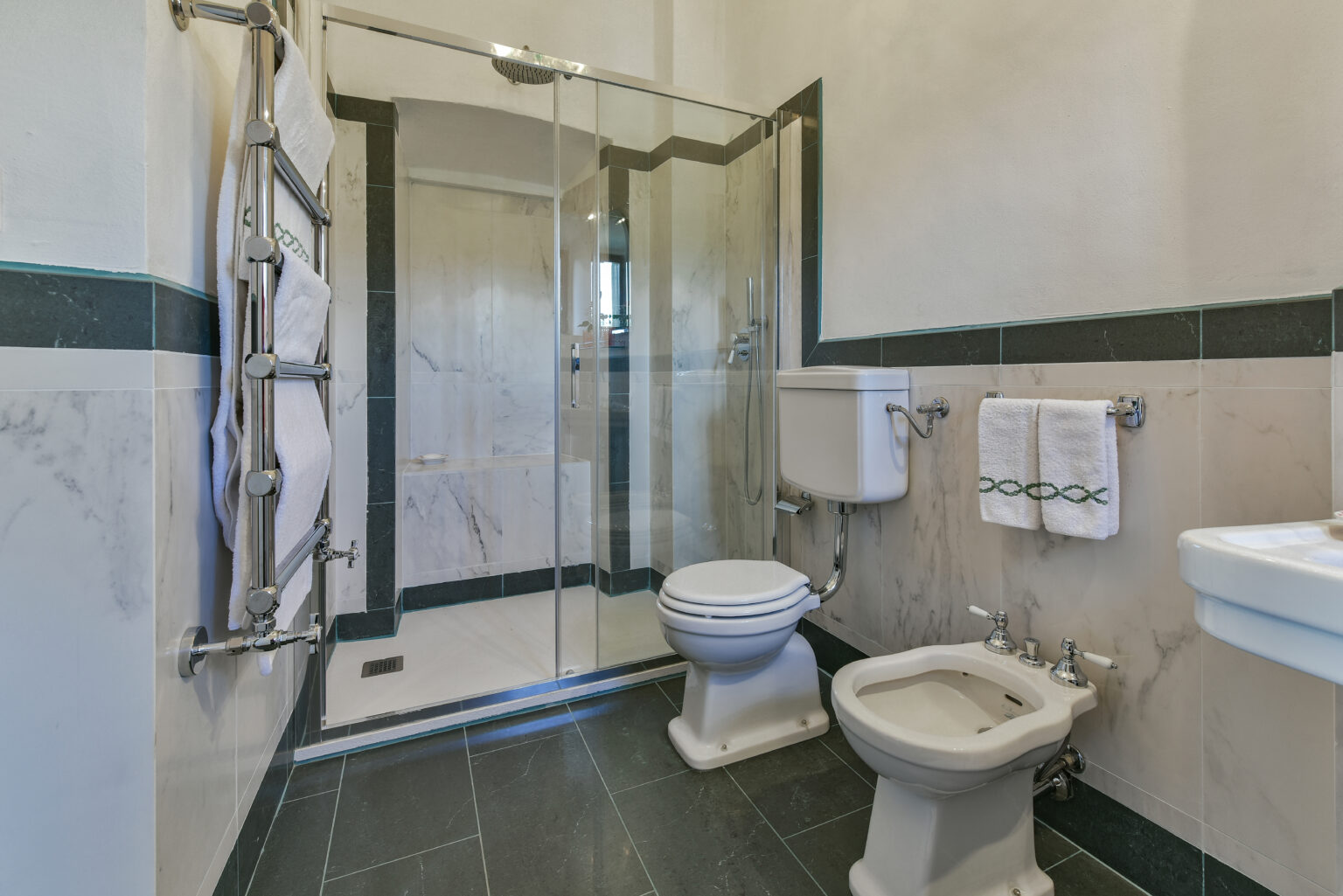 Modern bathroom featuring a glass shower, toilet, bidet, and sleek sink with marble accents and dark tile flooring. | Only on SopranoVillas