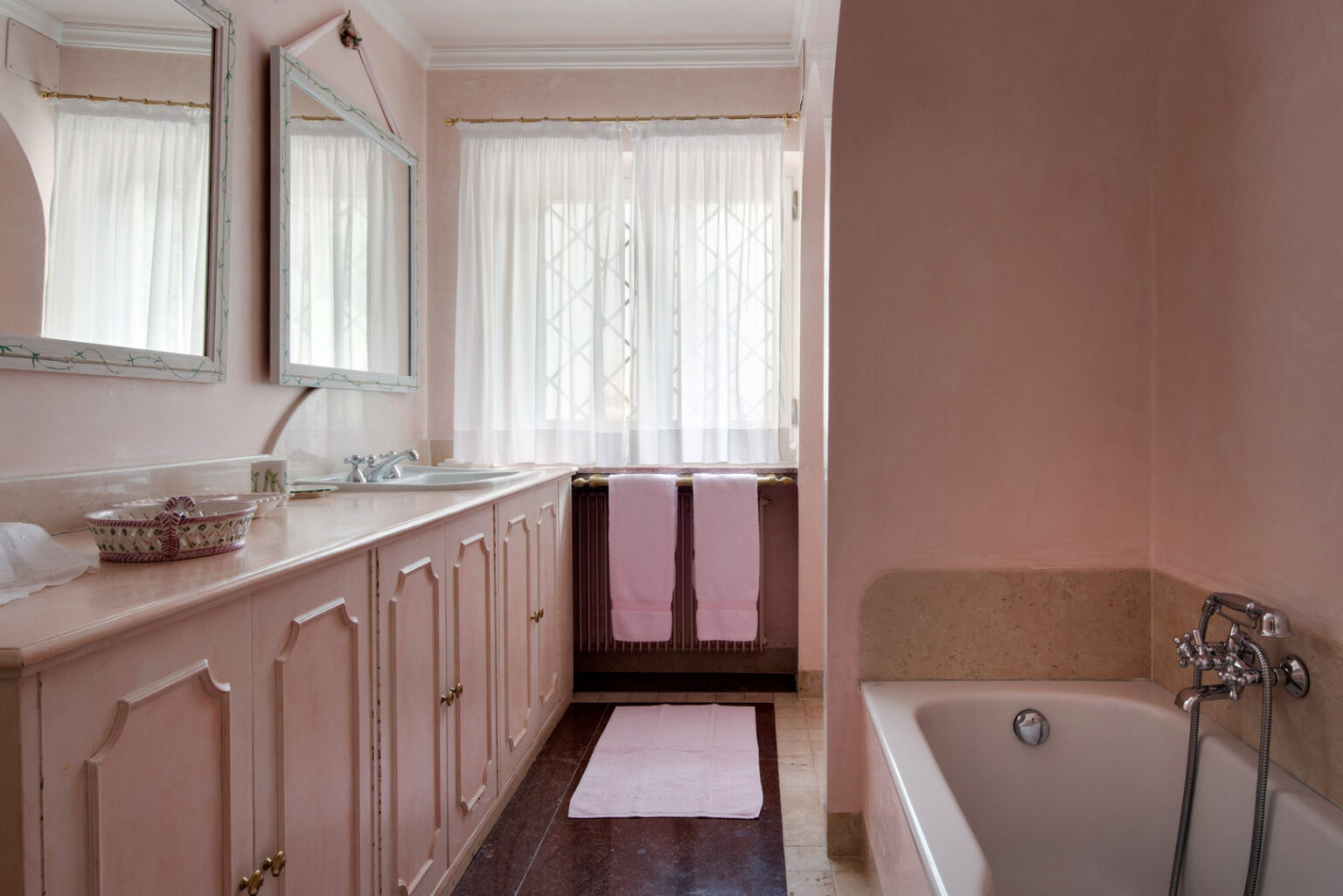 Bright and airy bathroom featuring pink walls, dual mirrors, a white bathtub, and a stylish vanity with storage. Natural light filters through sheer curtains, complemented by soft pink towels and a decorative bowl on the counter. | Only on SopranoVillas