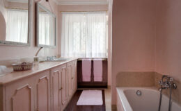 Bright and airy bathroom featuring pink walls, dual mirrors, a white bathtub, and a stylish vanity with storage. Natural light filters through sheer curtains, complemented by soft pink towels and a decorative bowl on the counter.