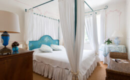Cozy bedroom featuring a blue canopy bed with floral pillows, white drapes, and soft lighting. The room includes a wooden dresser, a decorative lamp, and a small table with a vase of flowers, creating a serene and inviting atmosphere.