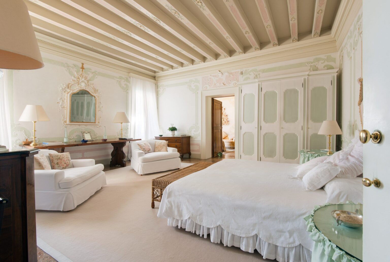 Spacious and elegantly designed bedroom featuring a white bed with a scalloped bed skirt, two comfortable white sofas with floral accents, and a wooden desk. The room boasts high, intricately detailed ceilings, soft pastel wall decor, and large windows allowing natural light to illuminate the space. A stylish mirror and decorative elements enhance the serene atmosphere. | Only on SopranoVillas