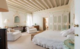 Spacious and elegantly designed bedroom featuring a white bed with a scalloped bed skirt, two comfortable white sofas with floral accents, and a wooden desk. The room boasts high, intricately detailed ceilings, soft pastel wall decor, and large windows allowing natural light to illuminate the space. A stylish mirror and decorative elements enhance the serene atmosphere.