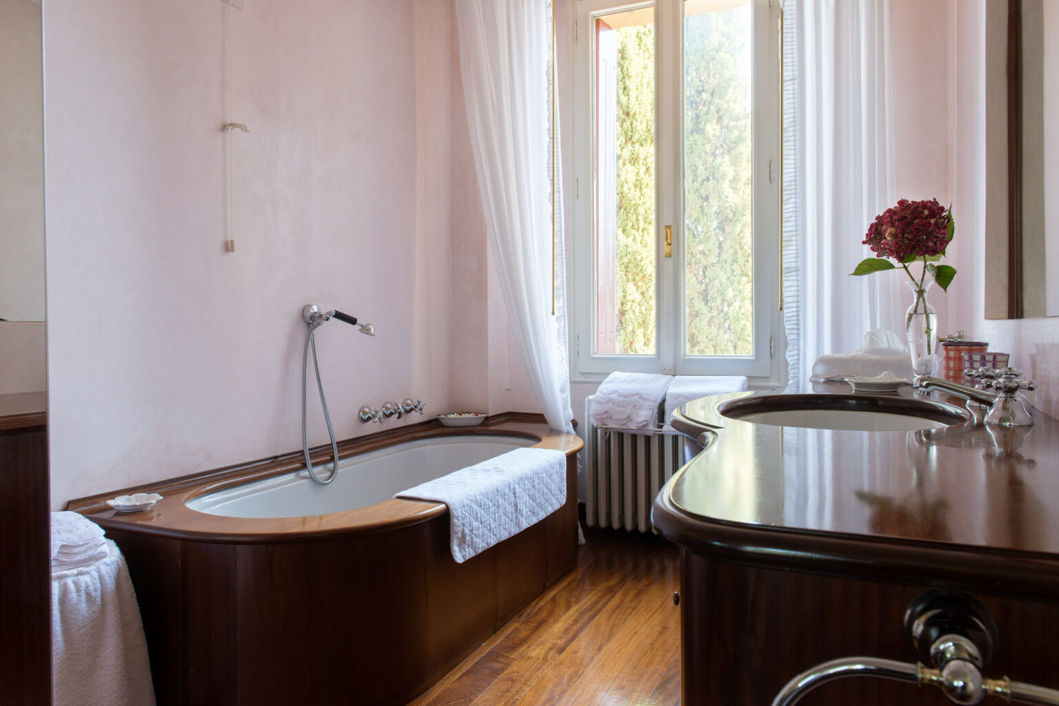Bright and elegant bathroom featuring a wooden bathtub, a stylish sink with modern fixtures, and large windows allowing natural light. The space is decorated with light pink walls, white curtains, and a floral arrangement, creating a serene and inviting atmosphere. Tidy towels and a radiator enhance the cozy feel of the room. | Only on SopranoVillas