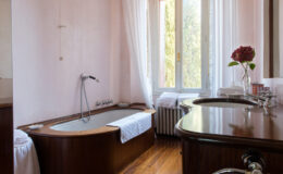 Bright and elegant bathroom featuring a wooden bathtub, a stylish sink with modern fixtures, and large windows allowing natural light. The space is decorated with light pink walls, white curtains, and a floral arrangement, creating a serene and inviting atmosphere. Tidy towels and a radiator enhance the cozy feel of the room.