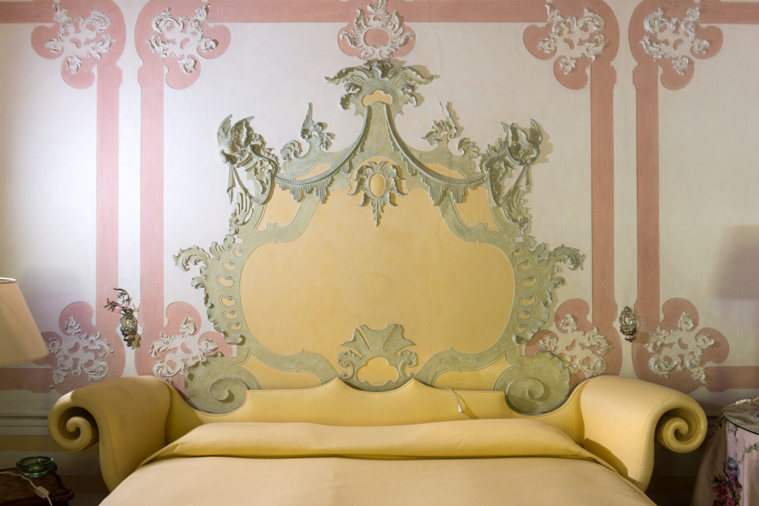 Elegant vintage bedroom featuring a decorative headboard in soft yellow and intricate wall detailing in pastel colors. The ornate design includes floral motifs and elegant curves, enhancing the room's classic aesthetic. A bedside lamp and table complement the serene atmosphere. | Only on SopranoVillas