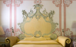 Elegant vintage bedroom featuring a decorative headboard in soft yellow and intricate wall detailing in pastel colors. The ornate design includes floral motifs and elegant curves, enhancing the room's classic aesthetic. A bedside lamp and table complement the serene atmosphere.