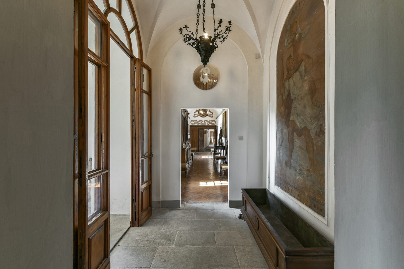 Elegant hallway featuring wooden doors, a vintage chandelier, and a decorative wall mural, leading to a spacious room. The stone floor adds to the classic ambiance of the interior design. | Only on SopranoVillas