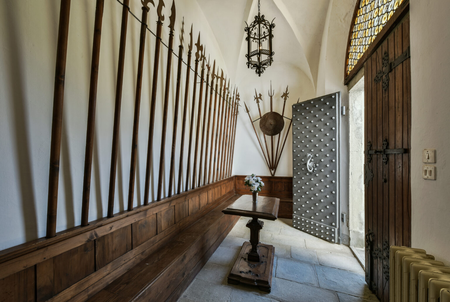 Historic entryway featuring medieval weapons displayed on the wall, a wooden bench, and an ornate metal door, illuminated by a decorative lantern. | Only on SopranoVillas