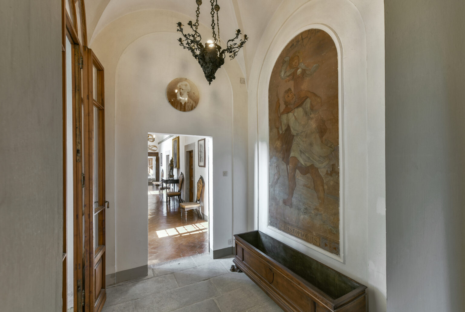 Elegant hallway featuring arched architecture, a decorative chandelier, and a wall mural, leading to a spacious living area with wooden furnishings. | Only on SopranoVillas