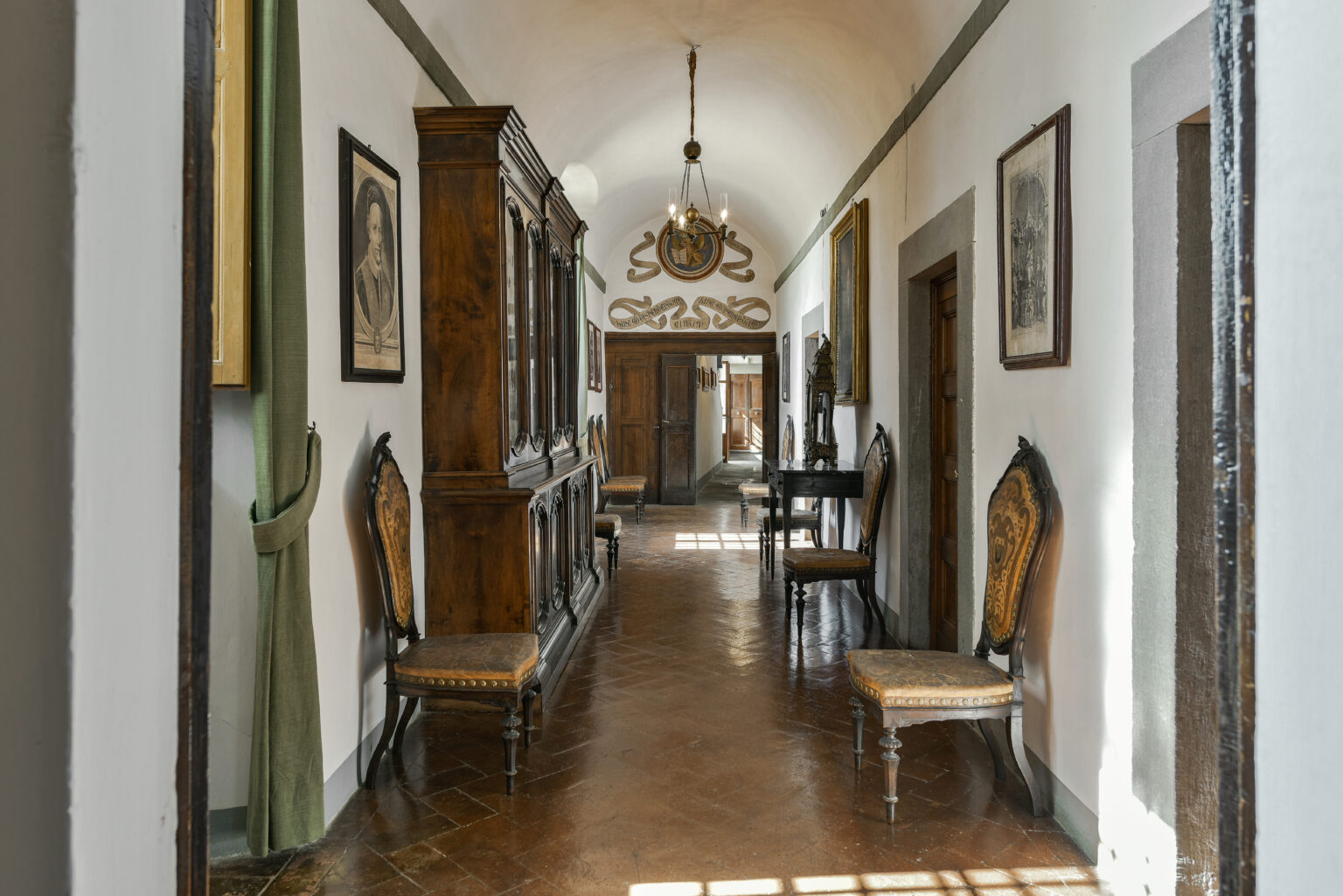 Interior view of a spacious hallway featuring wooden furniture, antique chairs, and framed artworks, illuminated by a chandelier, showcasing a blend of classic design and historical charm. | Only on SopranoVillas