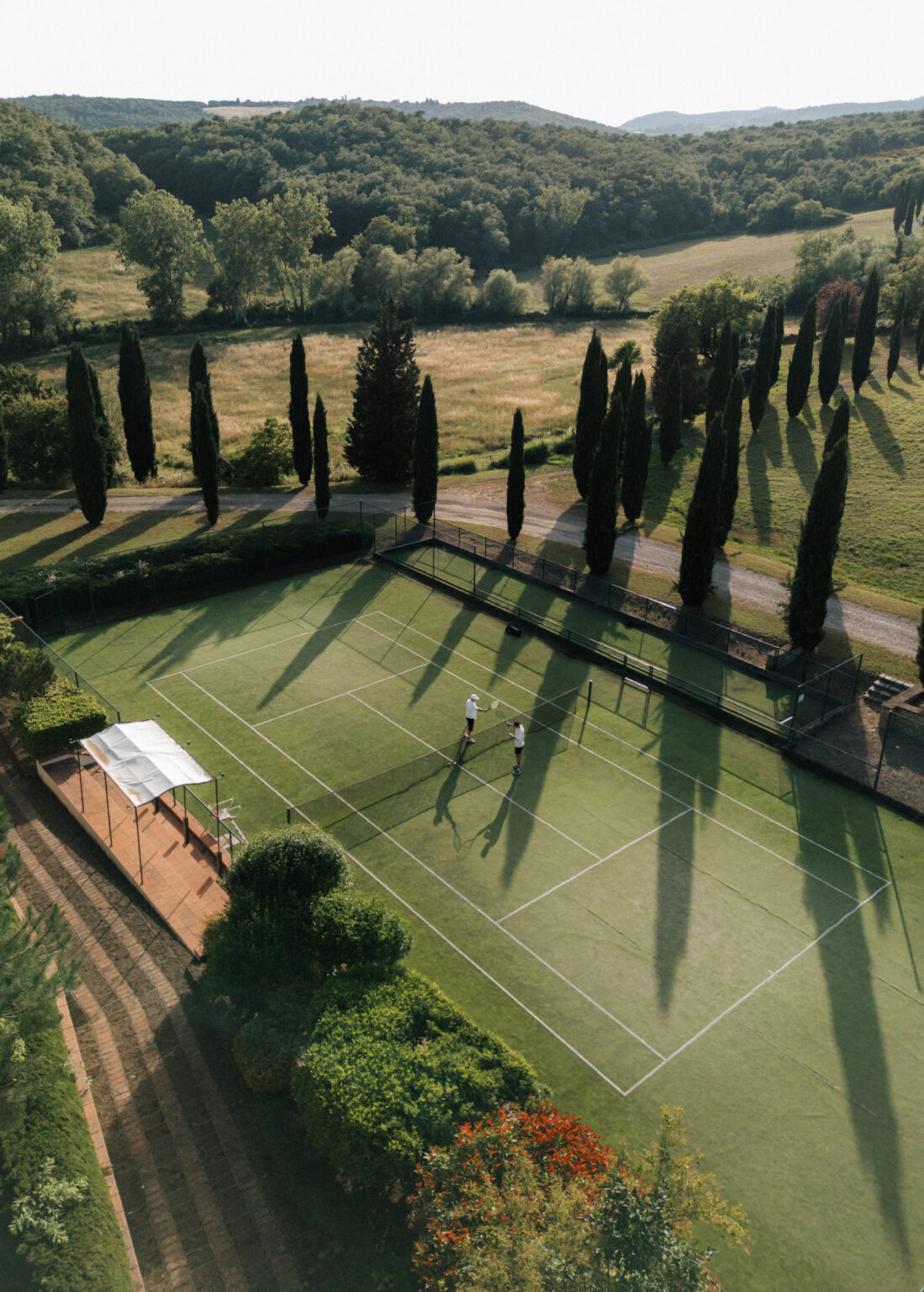 Aerial view of a tennis court surrounded by lush greenery and tall cypress trees, with two players engaged in a game. The court features well-defined lines and a shaded area for spectators, set against a backdrop of rolling hills and open fields. | Only on SopranoVillas