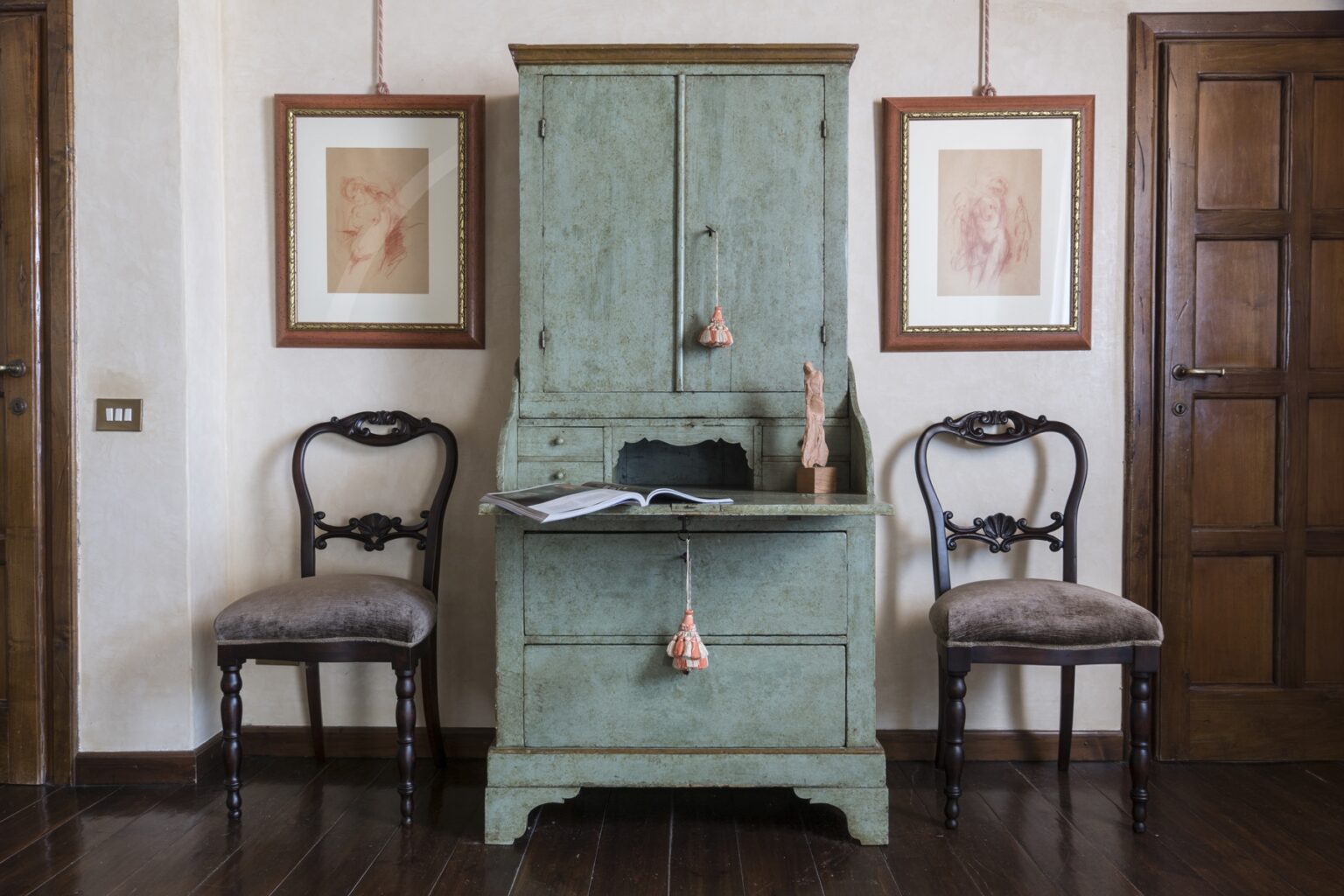 Vintage study area featuring a distressed green writing desk with decorative tassels, flanked by two elegant wooden chairs. The wall displays framed artworks, enhancing the cozy atmosphere of the room with warm wooden accents and a classic door. | Only on SopranoVillas