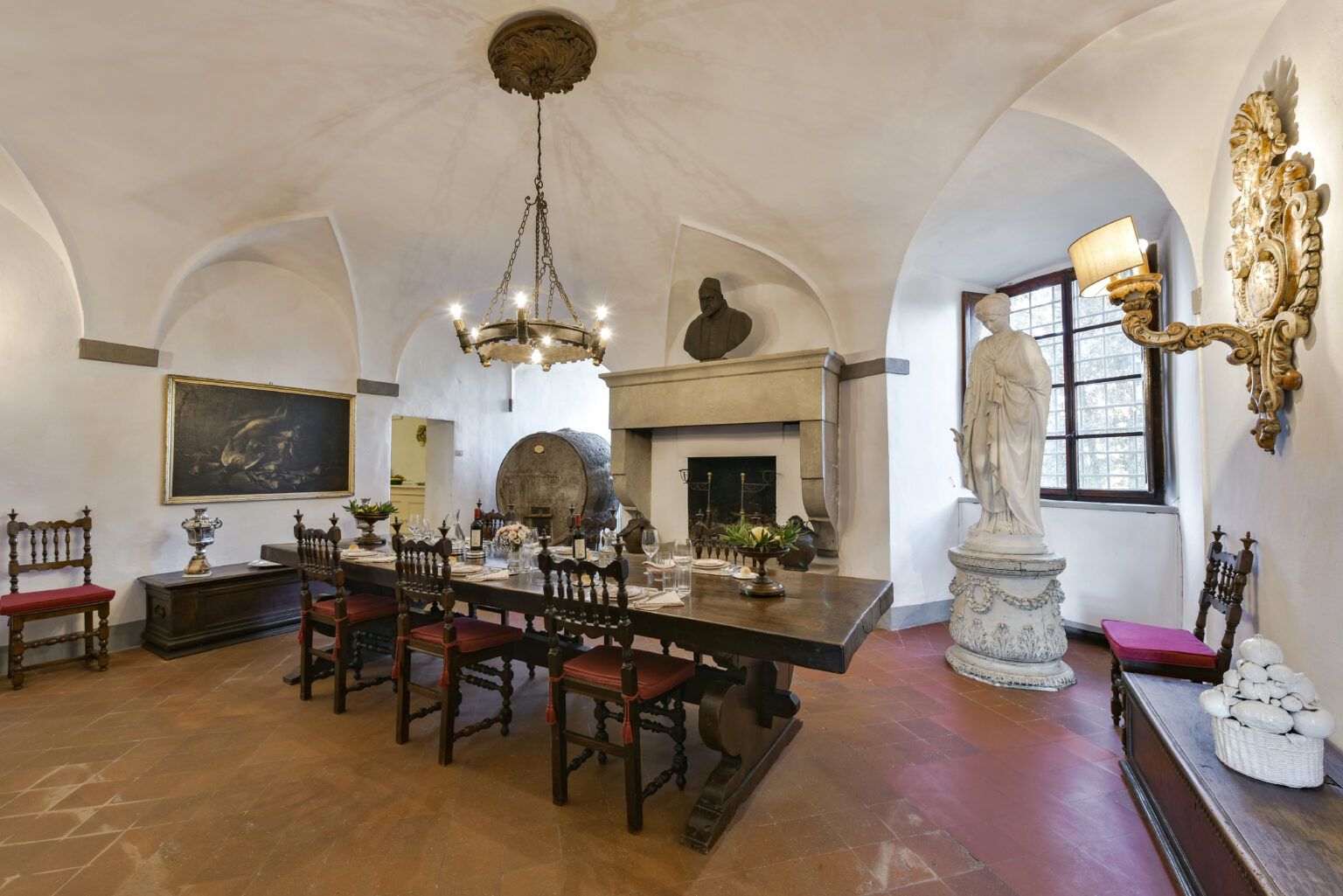 Historic dining room featuring a long wooden table surrounded by chairs, a stone fireplace, decorative wall art, and a statue. The room has arched ceilings and large windows, creating an elegant atmosphere. | Only on SopranoVillas