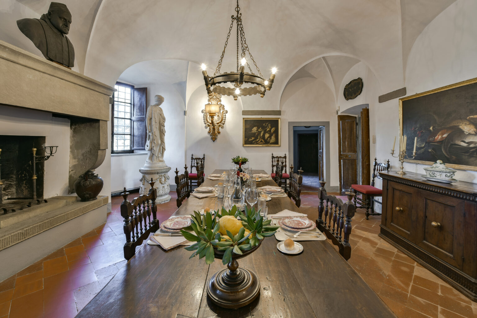 Historic dining room featuring a large wooden table set for a meal, surrounded by elegant chairs. A chandelier hangs from the vaulted ceiling, illuminating the space, while sculptures and artwork add to the room's classic charm. | Only on SopranoVillas